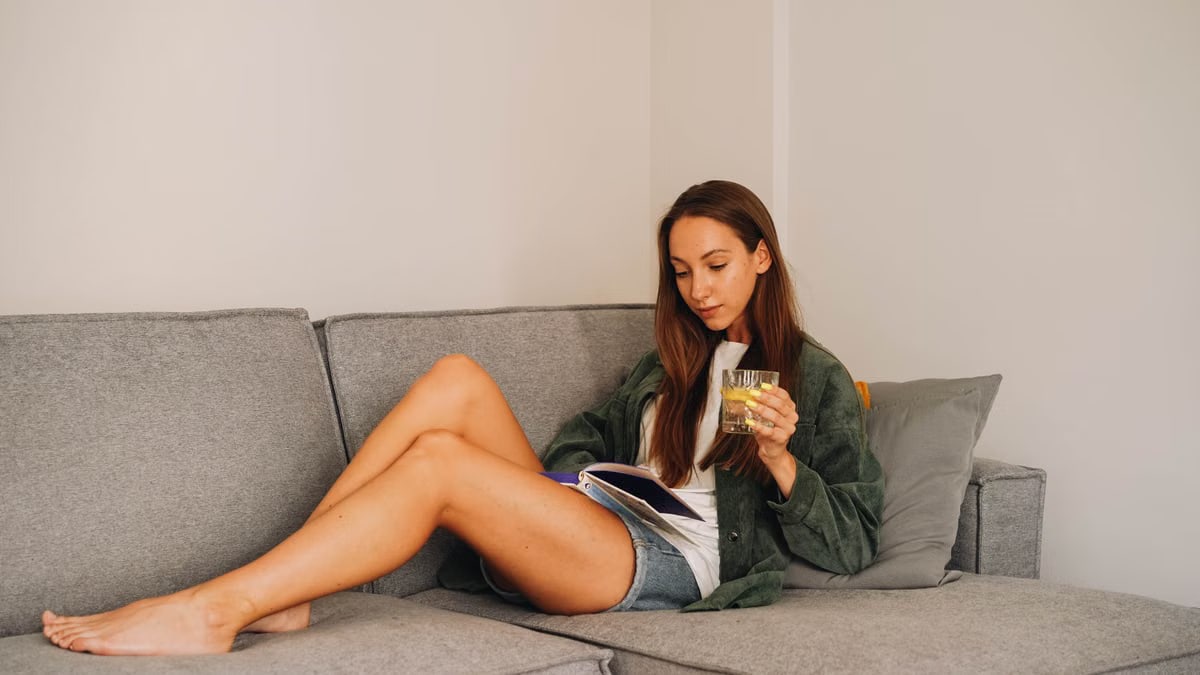 Woman reading a book on a grey sofa while holding a glass of water in a relaxed indoor setting.